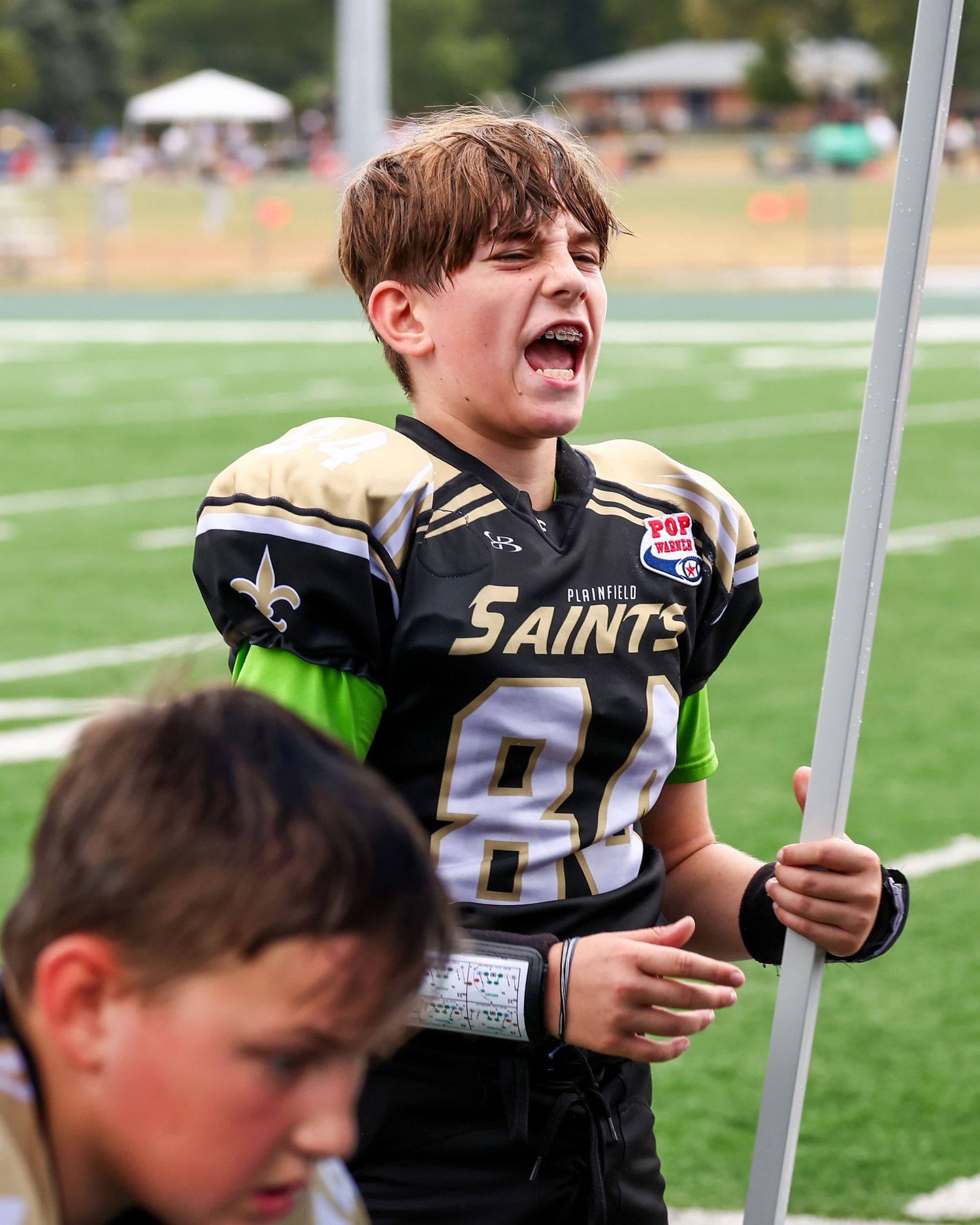 Young football player in black and gold Saints uniform cheers enthusiastically while holding a pole on the sidelines.