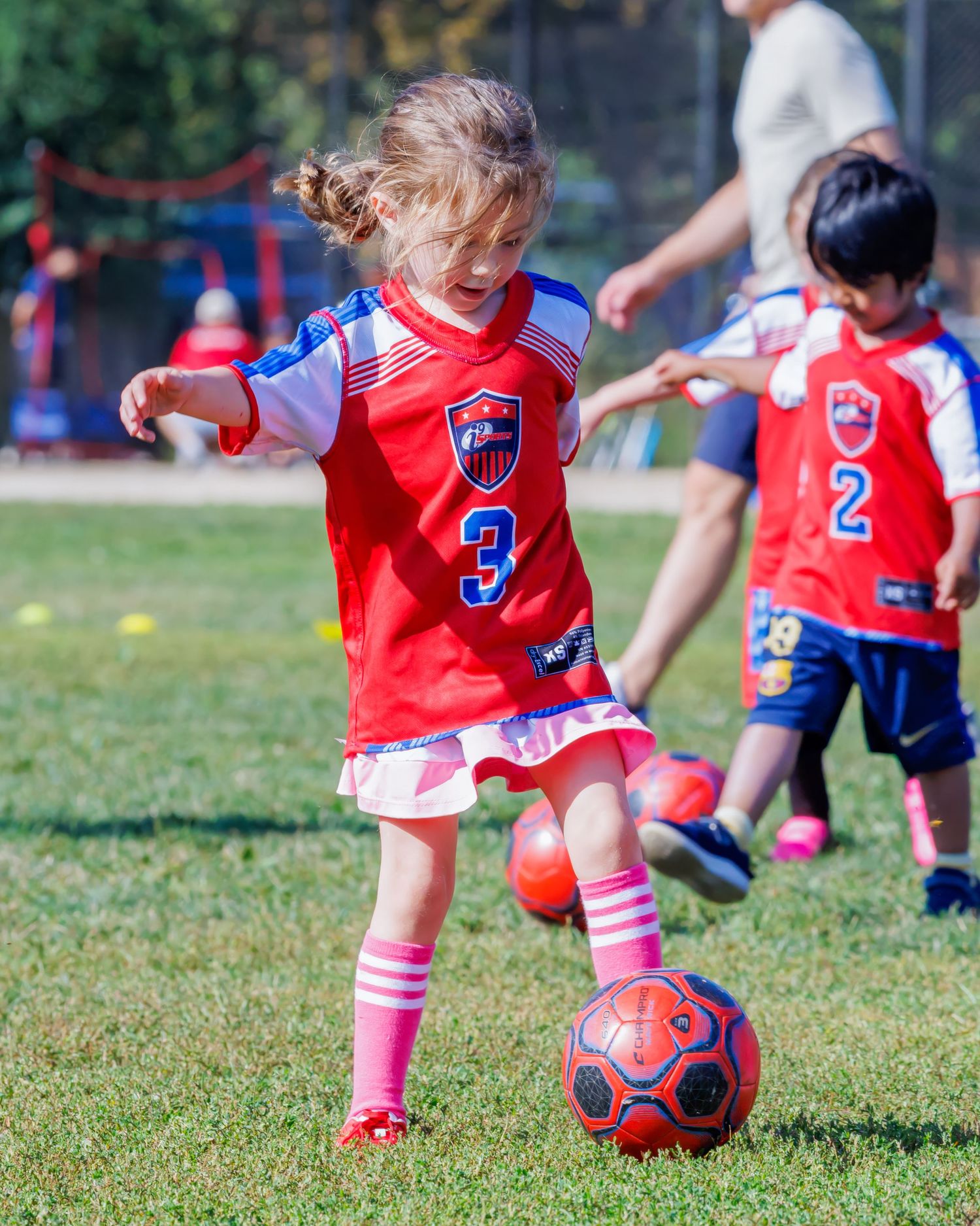 Young girl in red number 3 soccer jersey dribbling a ball during a youth soccer game on a green field.