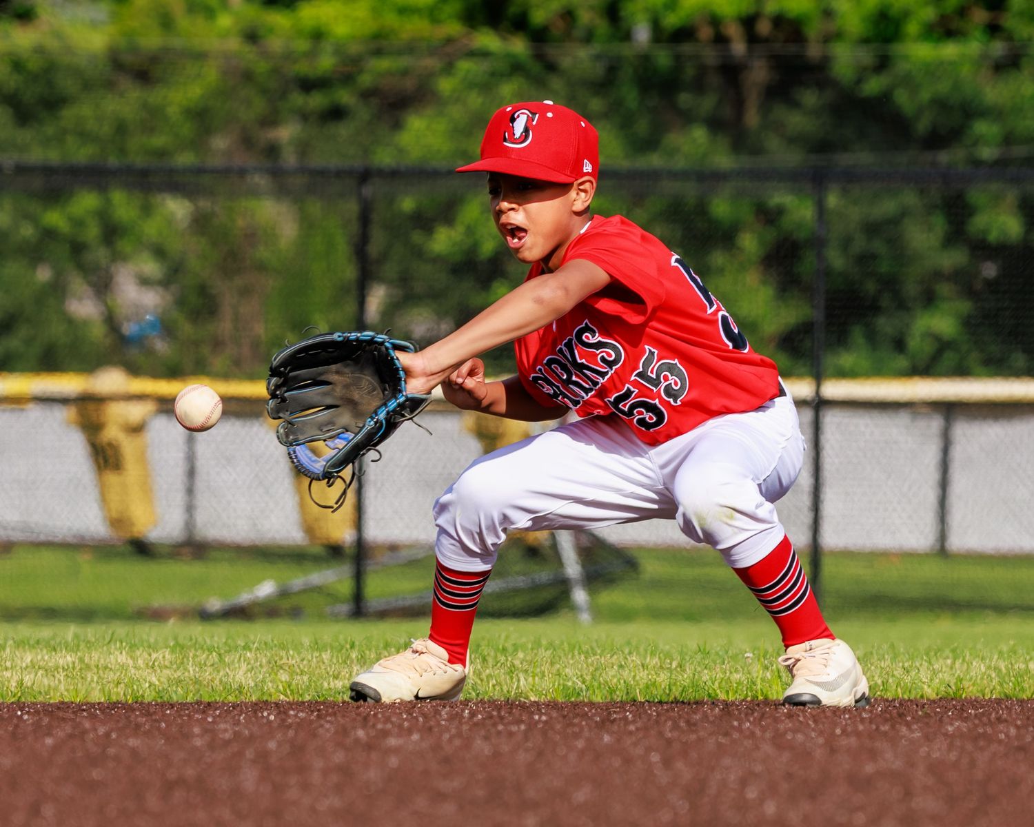 Young baseball player in red uniform fielding a ground ball in a crouched stance on the infield dirt.