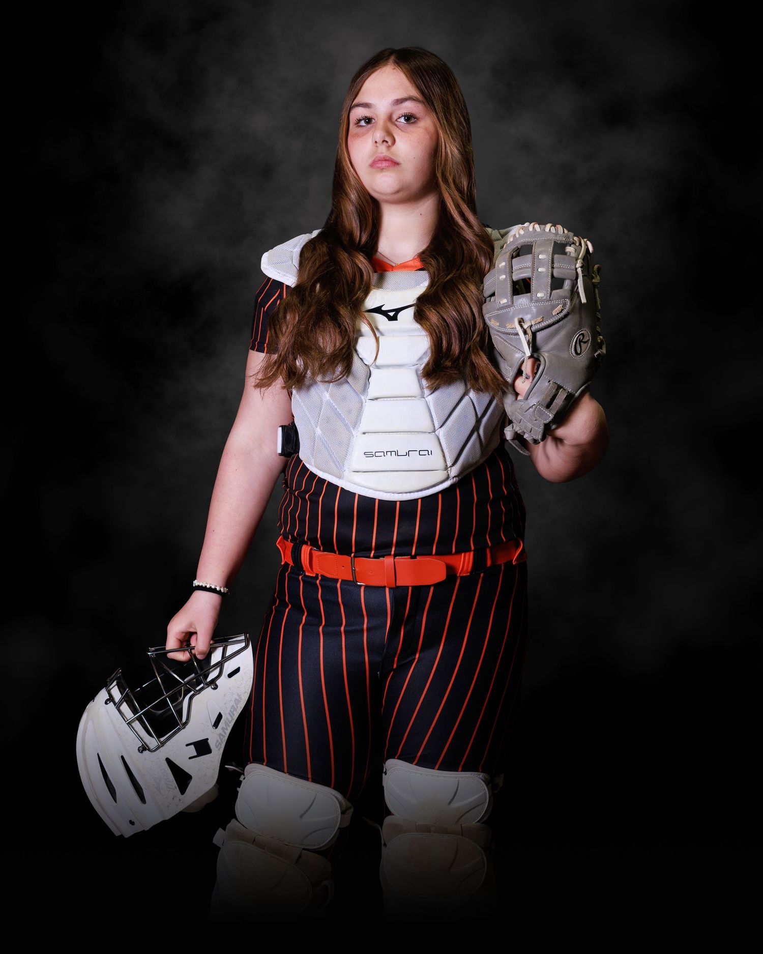 Young female softball catcher in uniform holding helmet and mitt, wearing chest protector and pinstripe pants.