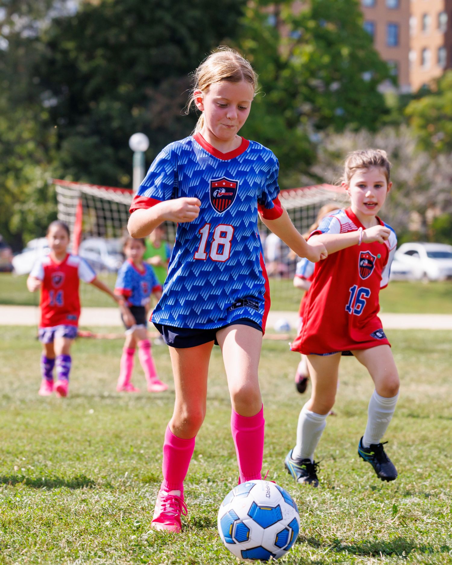 Young girls playing youth soccer match, player in blue jersey #18 dribbling ball while teammate in red #15 defends.