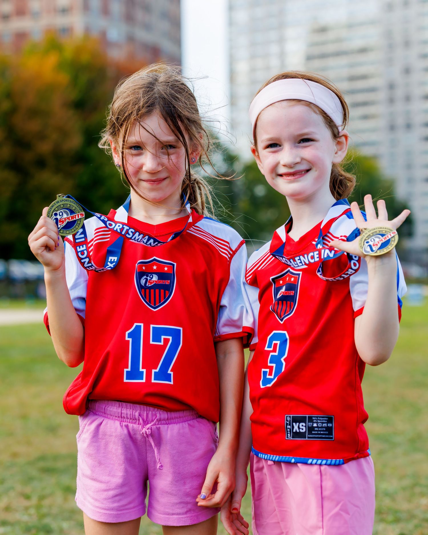 Two young girls in red soccer jerseys holding medals, wearing numbers 17 and 3, smiling outdoors after a game.