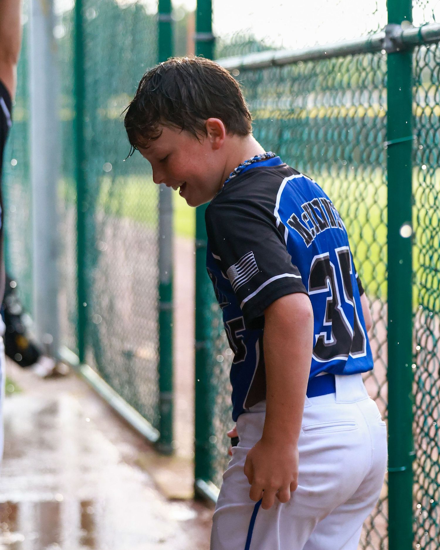 Young player in baseball dugout watching game through chain link fence.