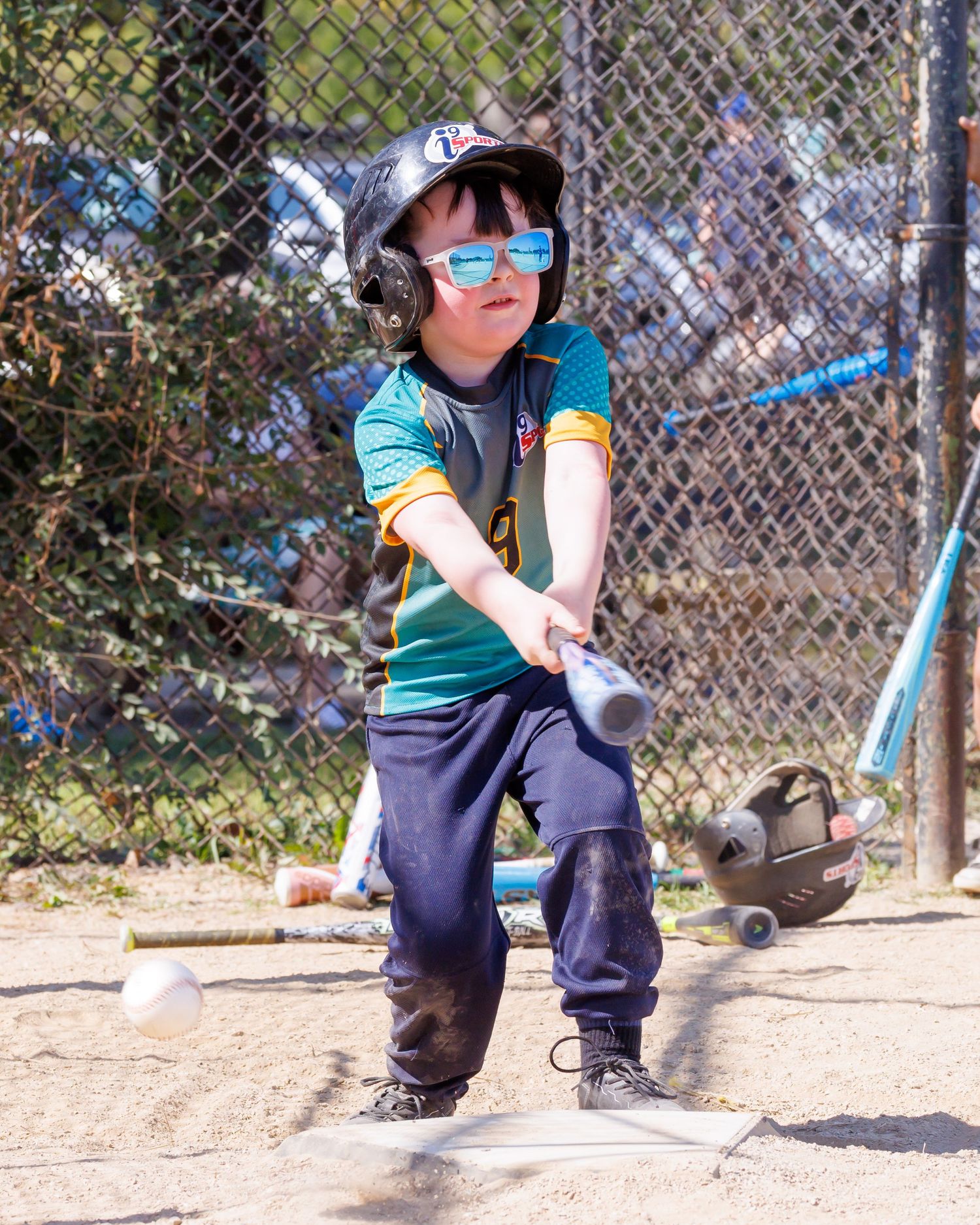 Young baseball player in teal jersey and sunglasses takes batting stance at home plate.
