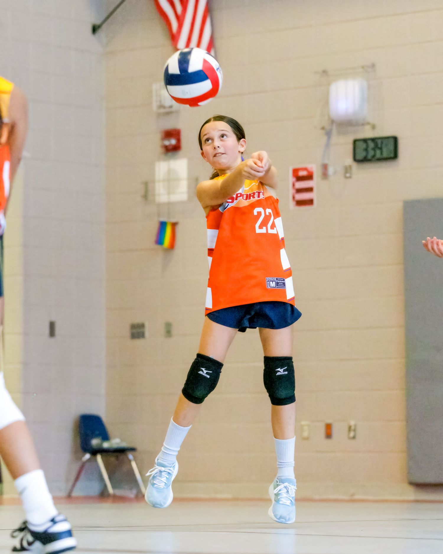 Youth volleyball players in orange jerseys practicing receiving drills in a gymnasium.