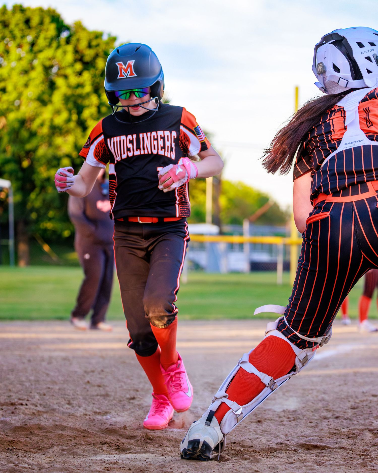 Mudslingers softball player in pink cleats runs base as catcher in orange pinstripes defends during game.
