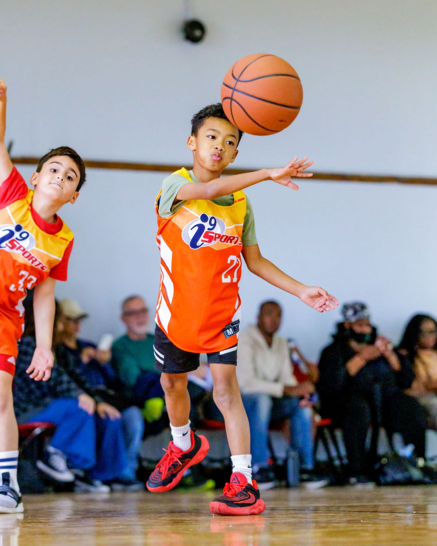 Young basketball players in orange jerseys dive for loose balls during an energetic youth league game on a wooden court.