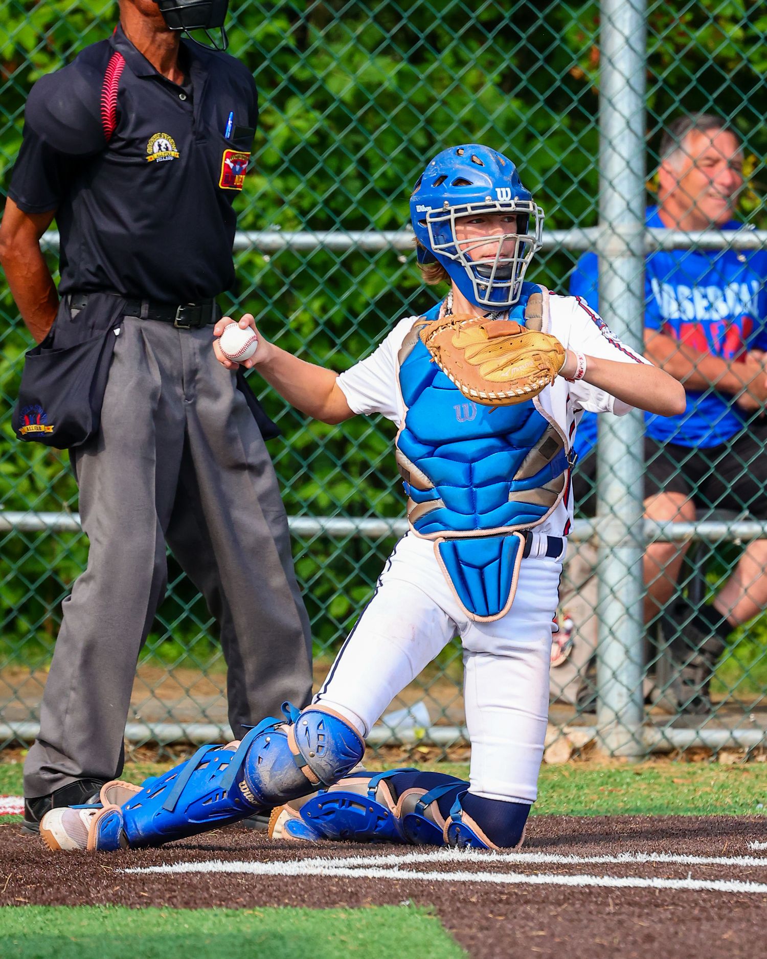 Baseball catcher in blue gear blocks home plate during an intense play at a youth baseball game.
