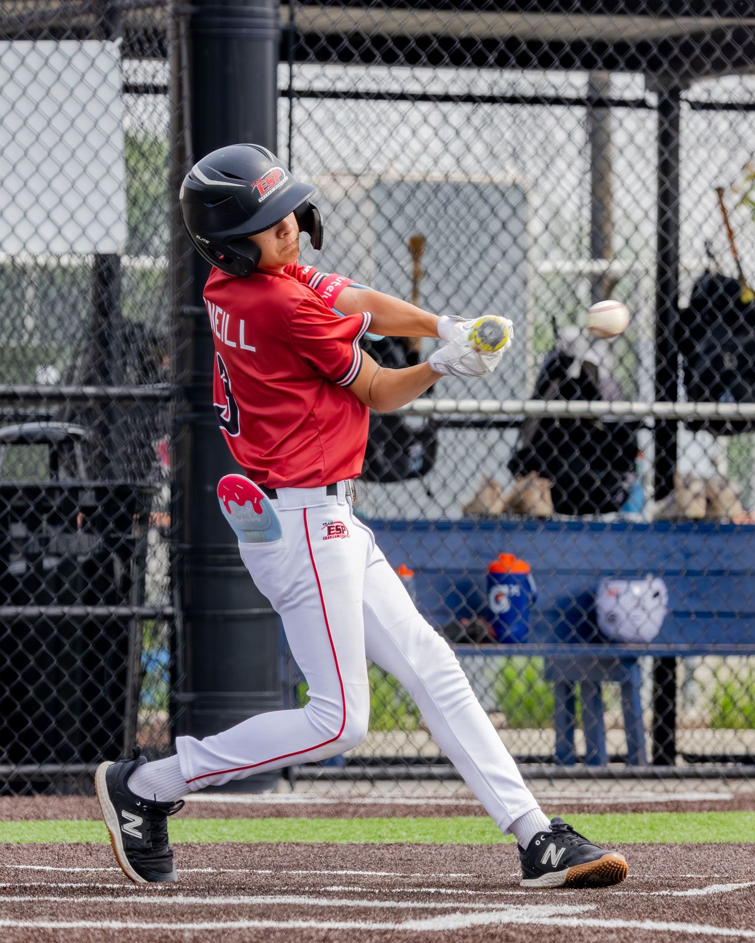 Baseball batter in red jersey and helmet swings at pitch during game at outdoor batting cage.