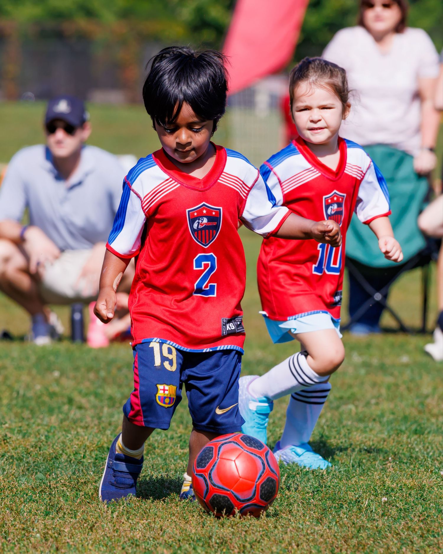 Young soccer players in red and blue uniforms practicing ball control drills on a sunny grass field.