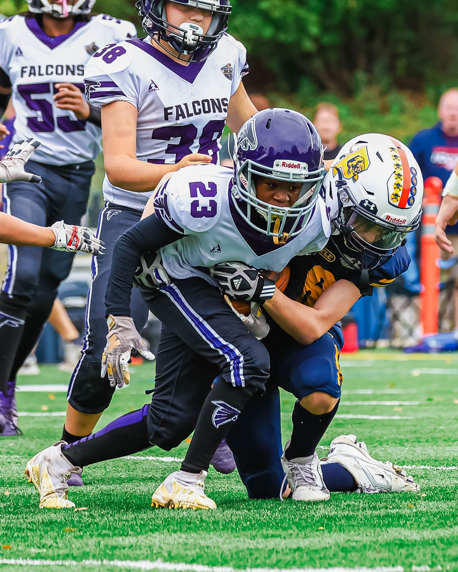 Youth football player #23 in purple Falcons jersey carries the ball while being tackled during a youth league game.