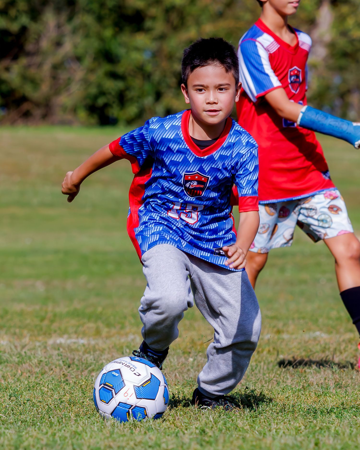 Young boy in blue soccer jersey dribbles ball during youth soccer match on green grass field.