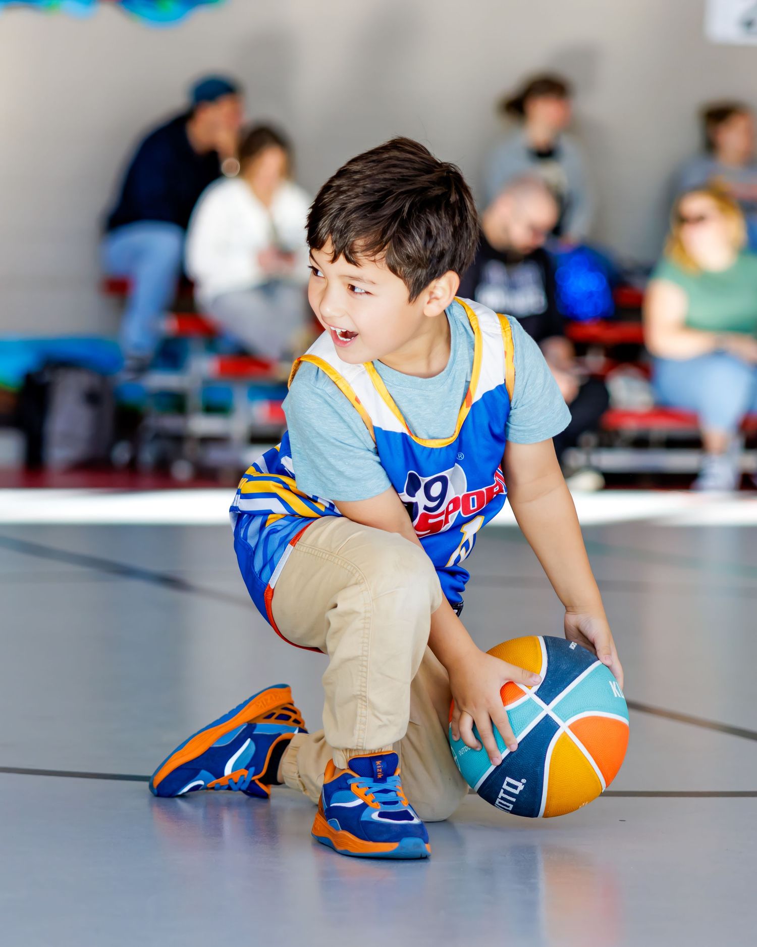 Young boy in blue jersey dribbles an orange basketball on an indoor court during a youth basketball game.