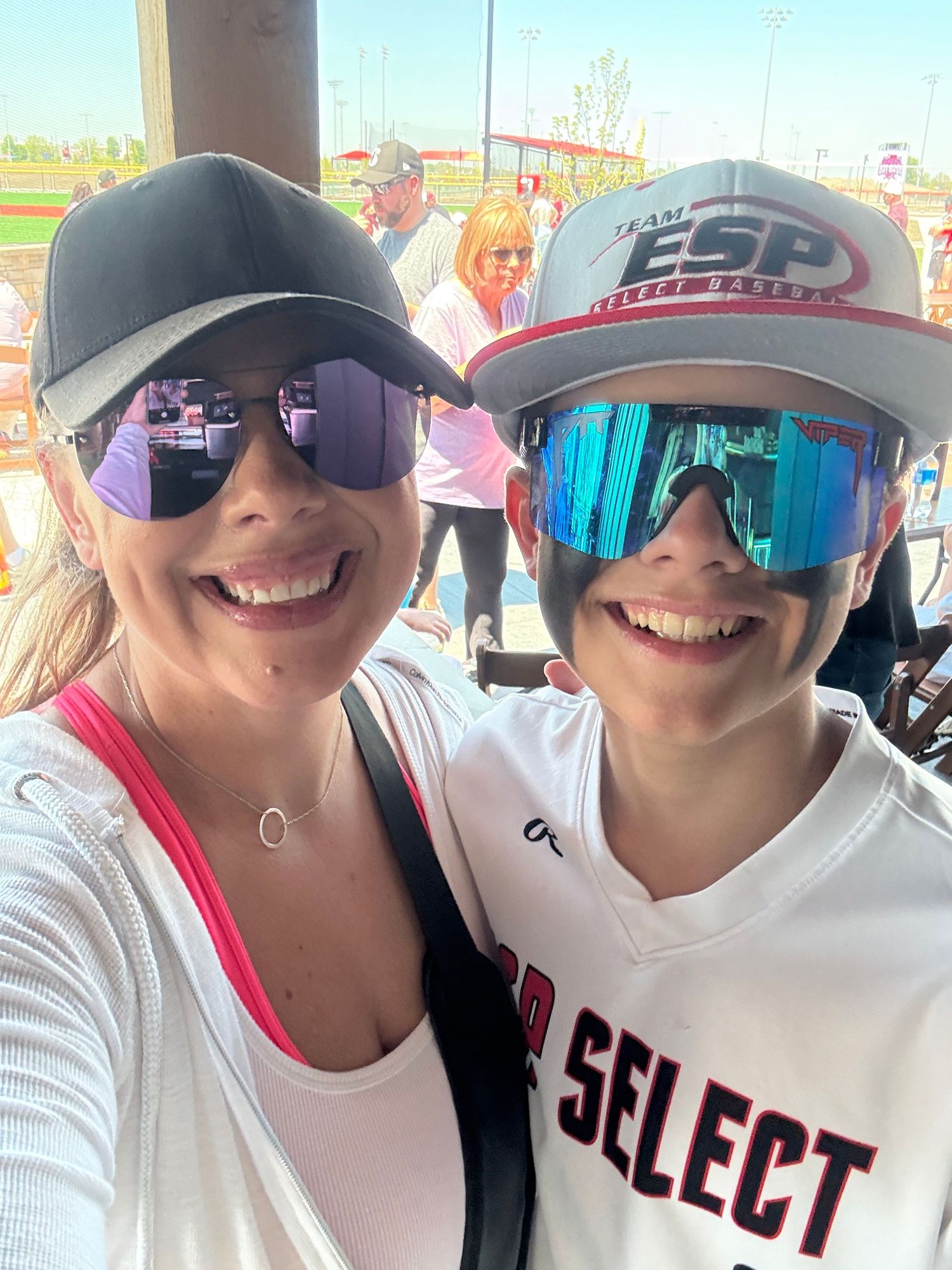 Two people wearing baseball caps and sunglasses posing at a sports venue.