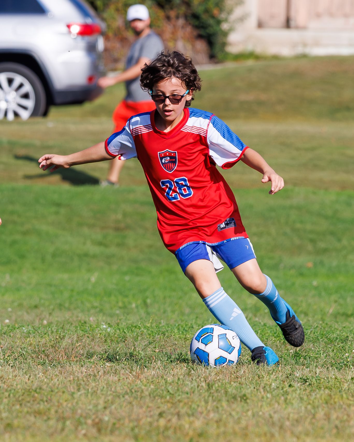 Young soccer player #26 in red and blue uniform kicks ball on grass field during youth league game.