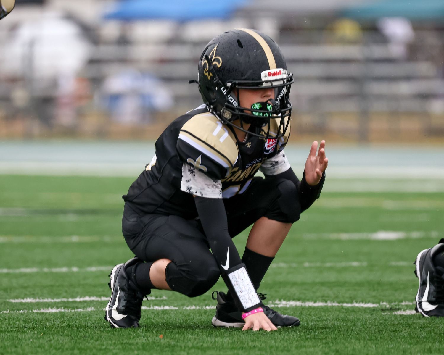 Youth football player in black Saints uniform crouching on field during game.