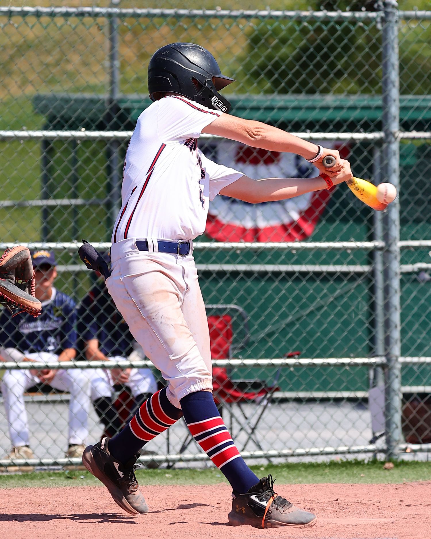 Baseball batter in white uniform follows through on swing during game with chain link fence visible in background.