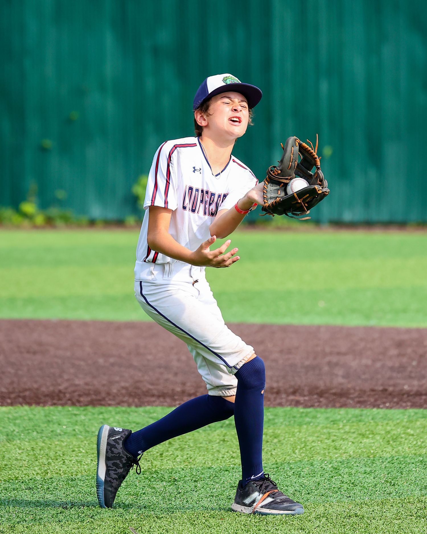 Young baseball player in white uniform and navy cap catches a pop fly on grass field during training.