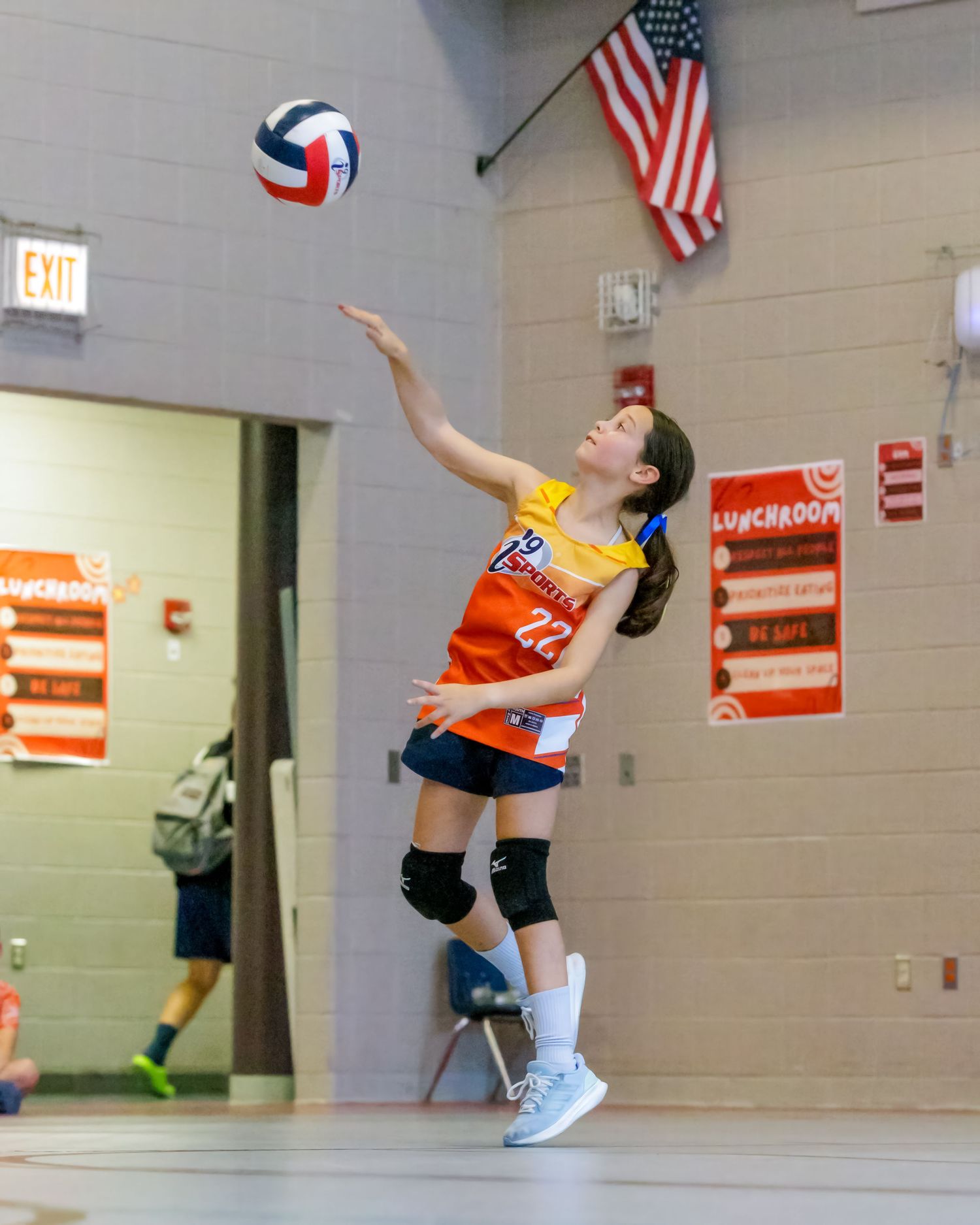 Volleyball player in orange jersey leaps high to serve ball during indoor match with American flags on wall.