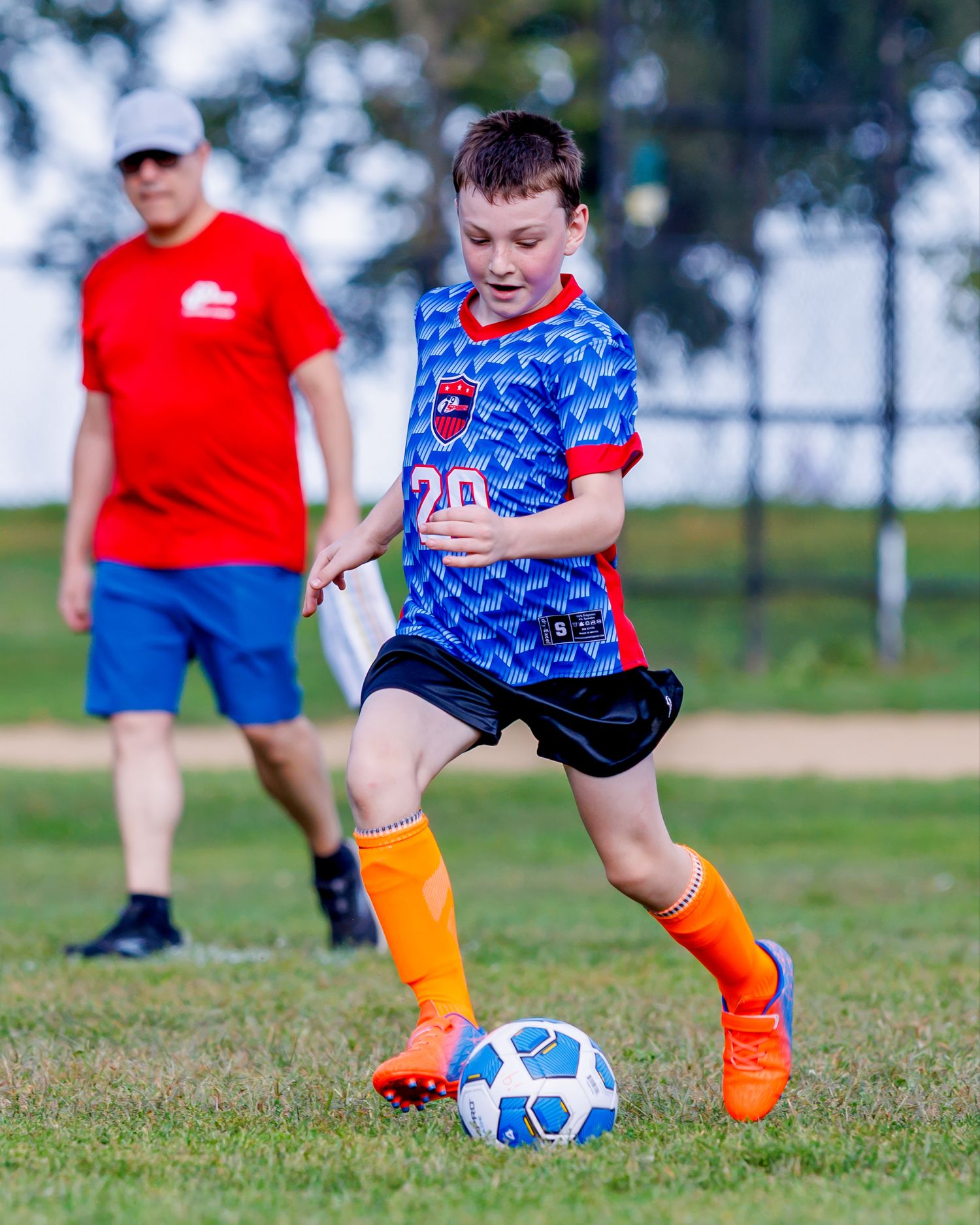 Young soccer player in blue and orange uniform practicing ball control drills on a grassy field.