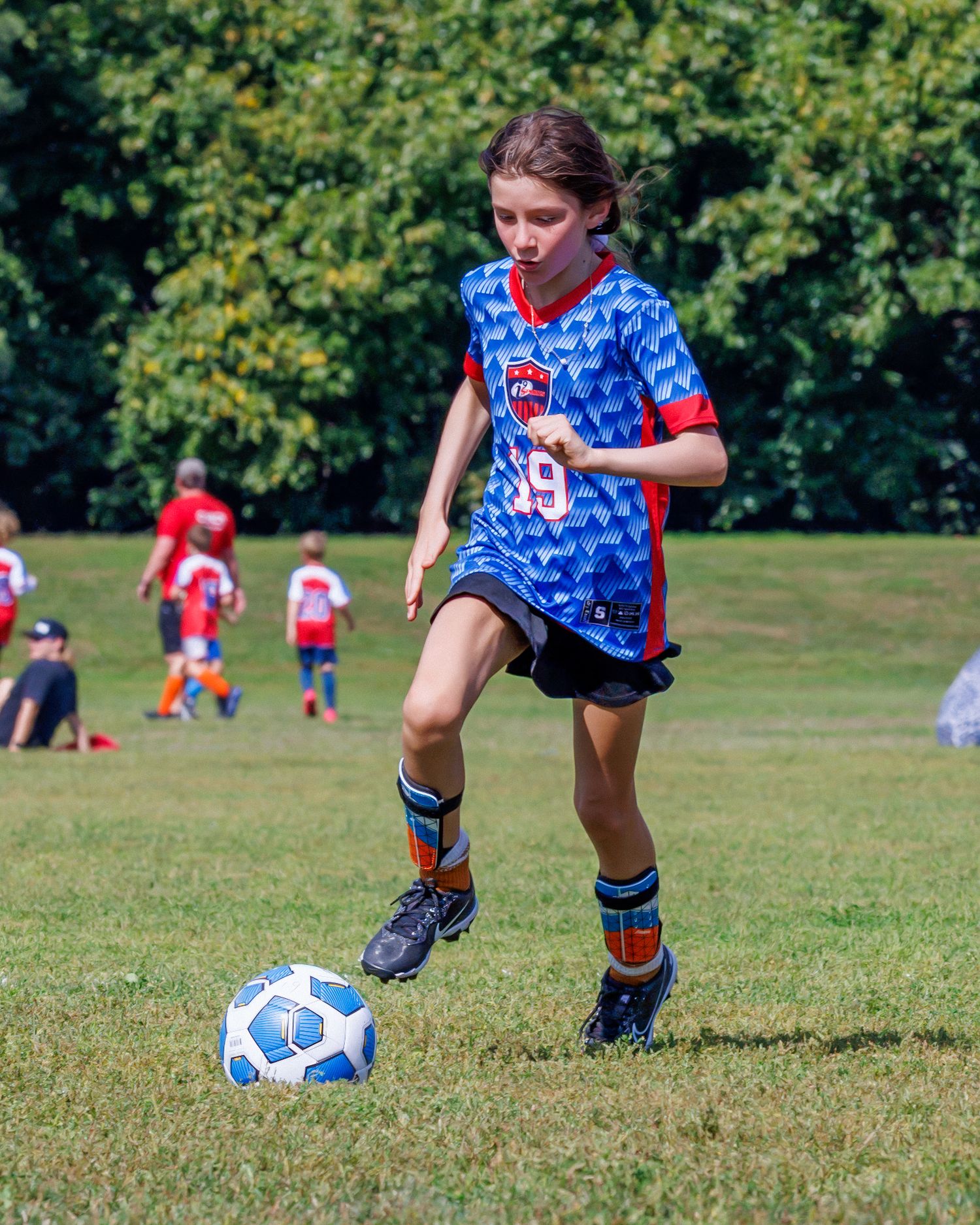 Young girl in blue soccer jersey dribbling a ball on a green grass field during a youth soccer game.