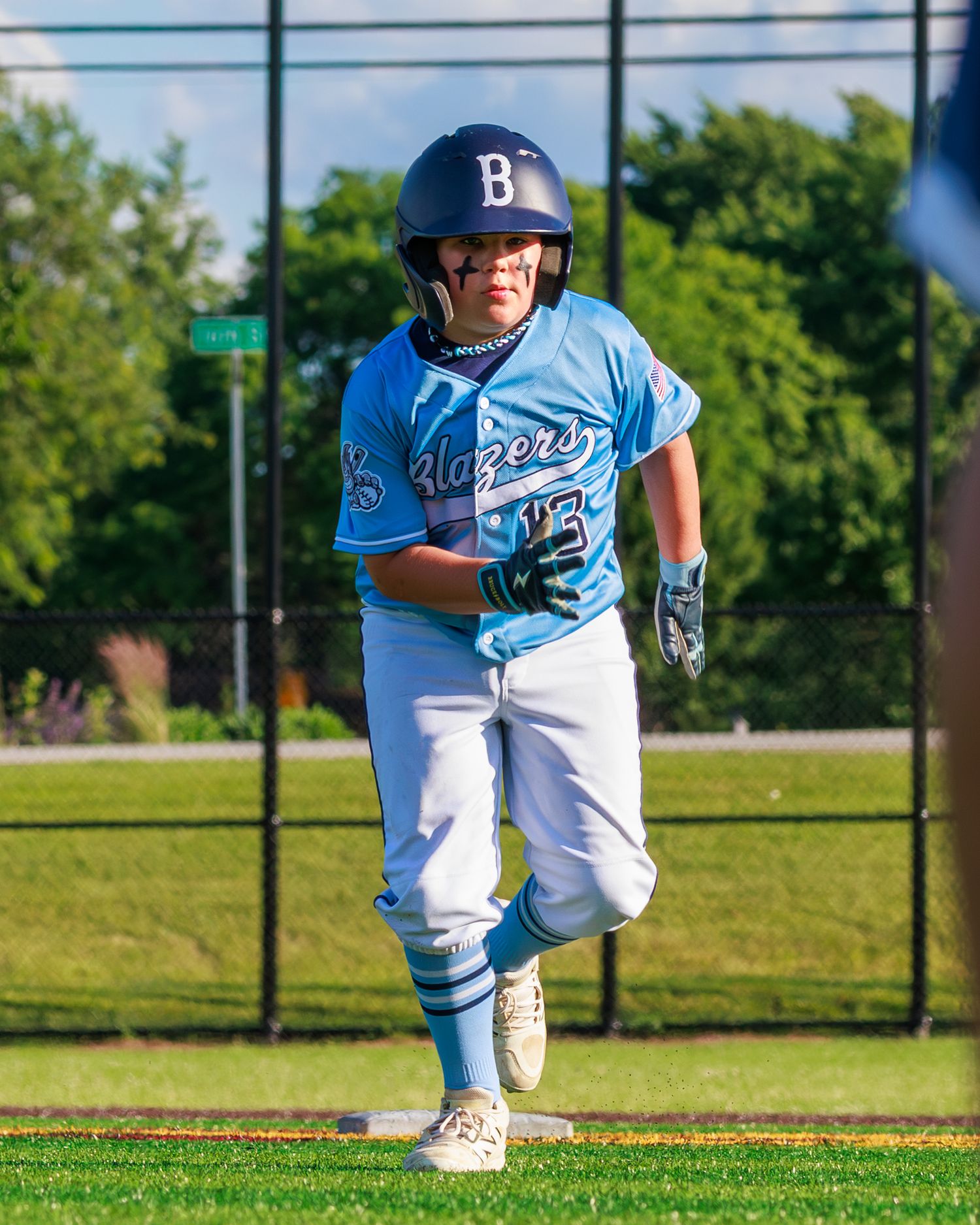 Young baseball player in blue Blazers uniform and helmet runs the bases during a youth league game on a sunny day.