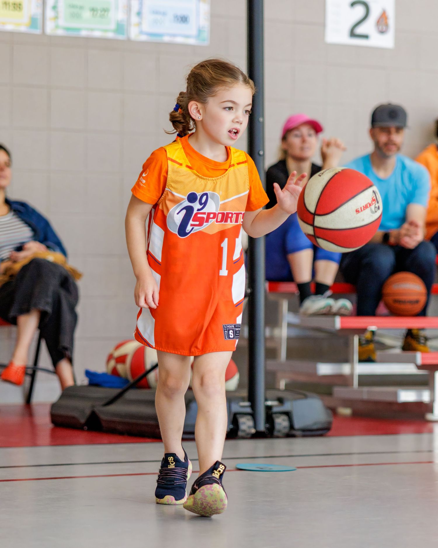 Young girl in orange iSports jersey dribbling a basketball during a youth indoor basketball game.