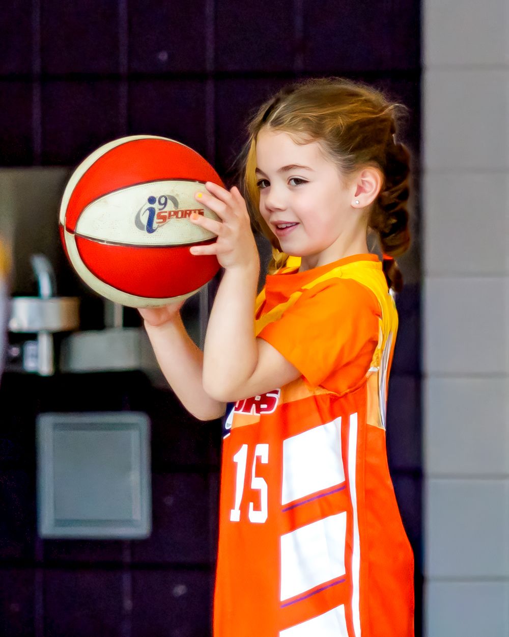 A player in an orange jersey practices shooting with a red and white basketball in a gym.