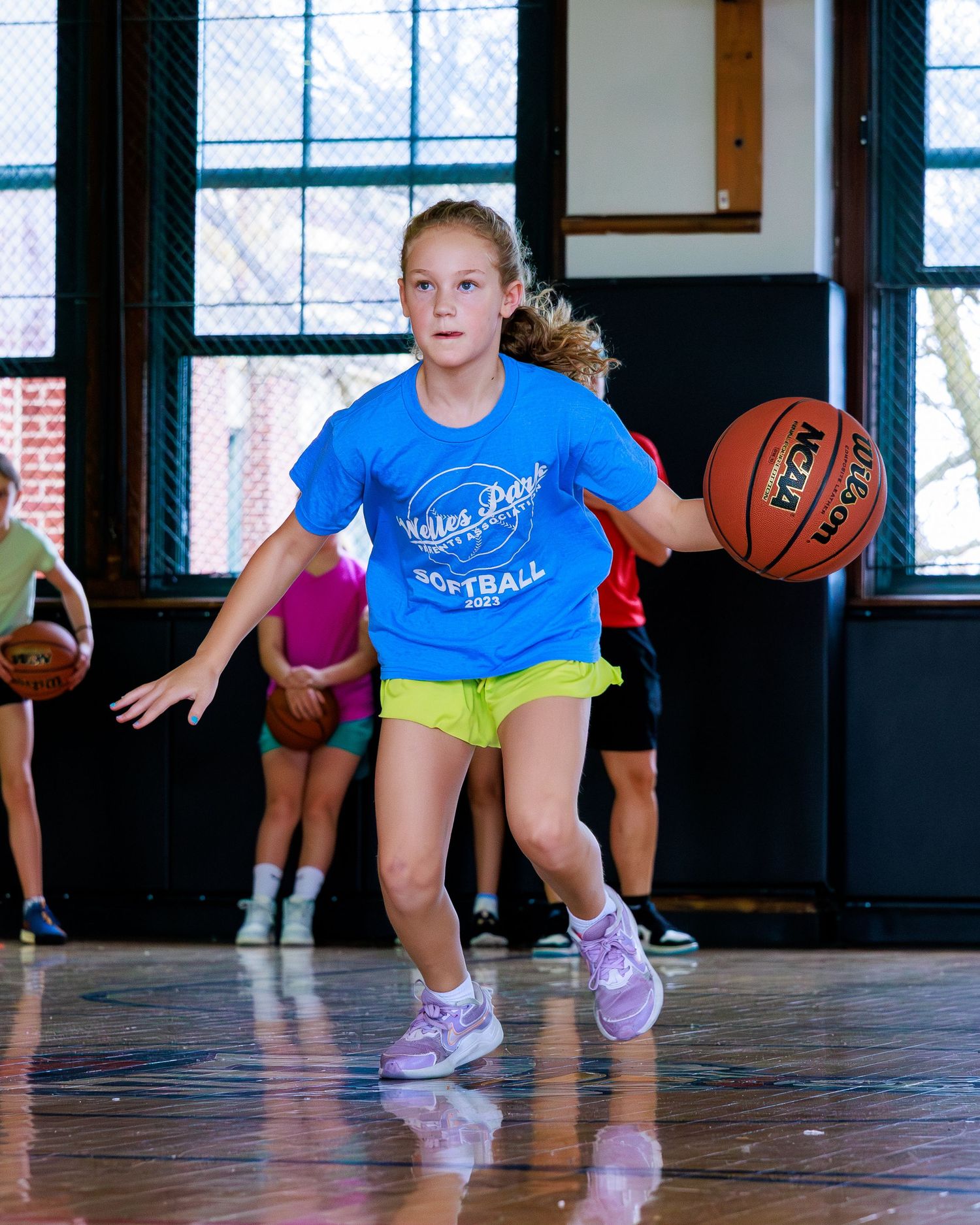 Young basketball players in blue shirts practice defensive stance drills on a wooden gym floor.