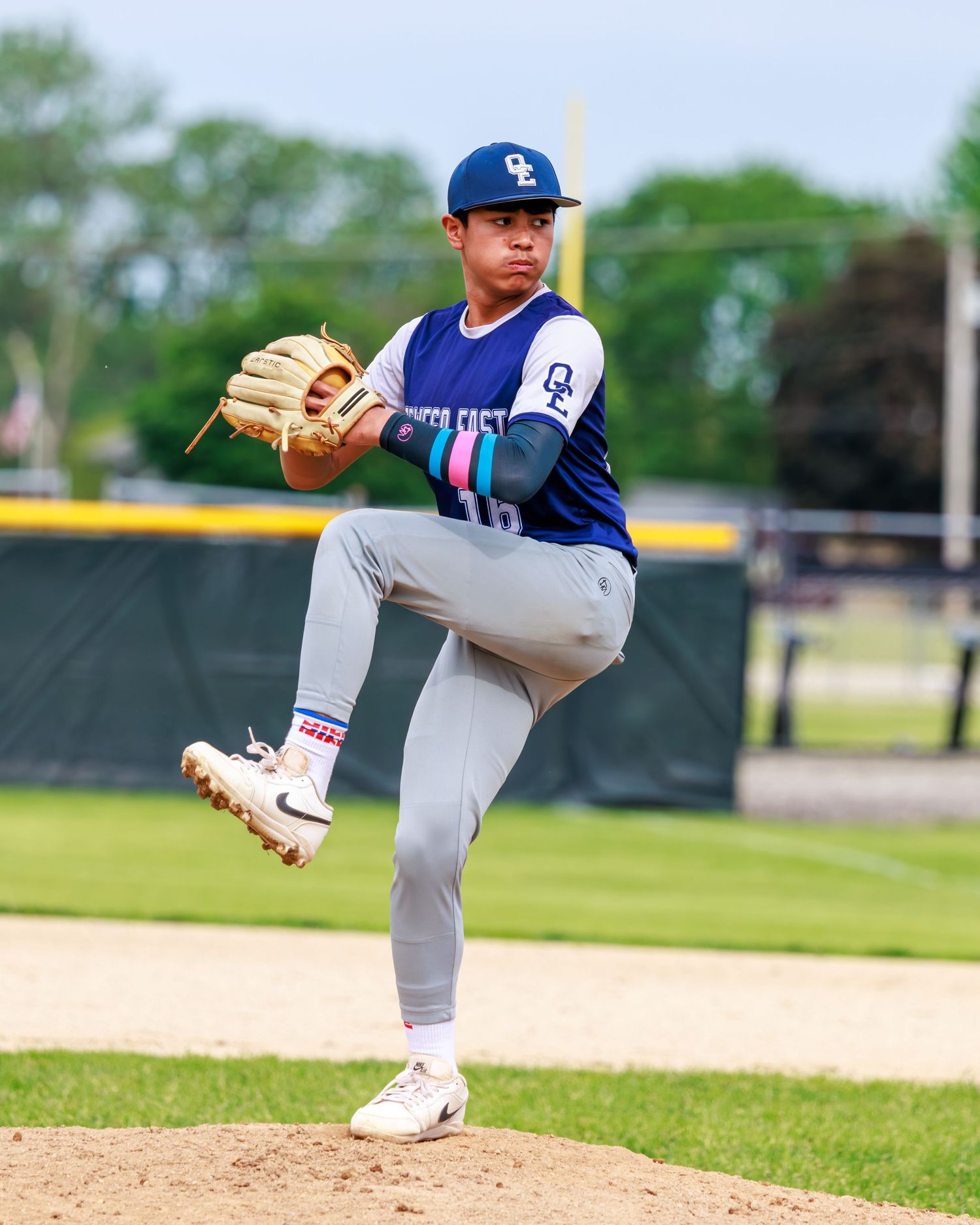 A youth baseball pitcher in a blue cap and gray uniform winds up to throw on a sunny outdoor mound.