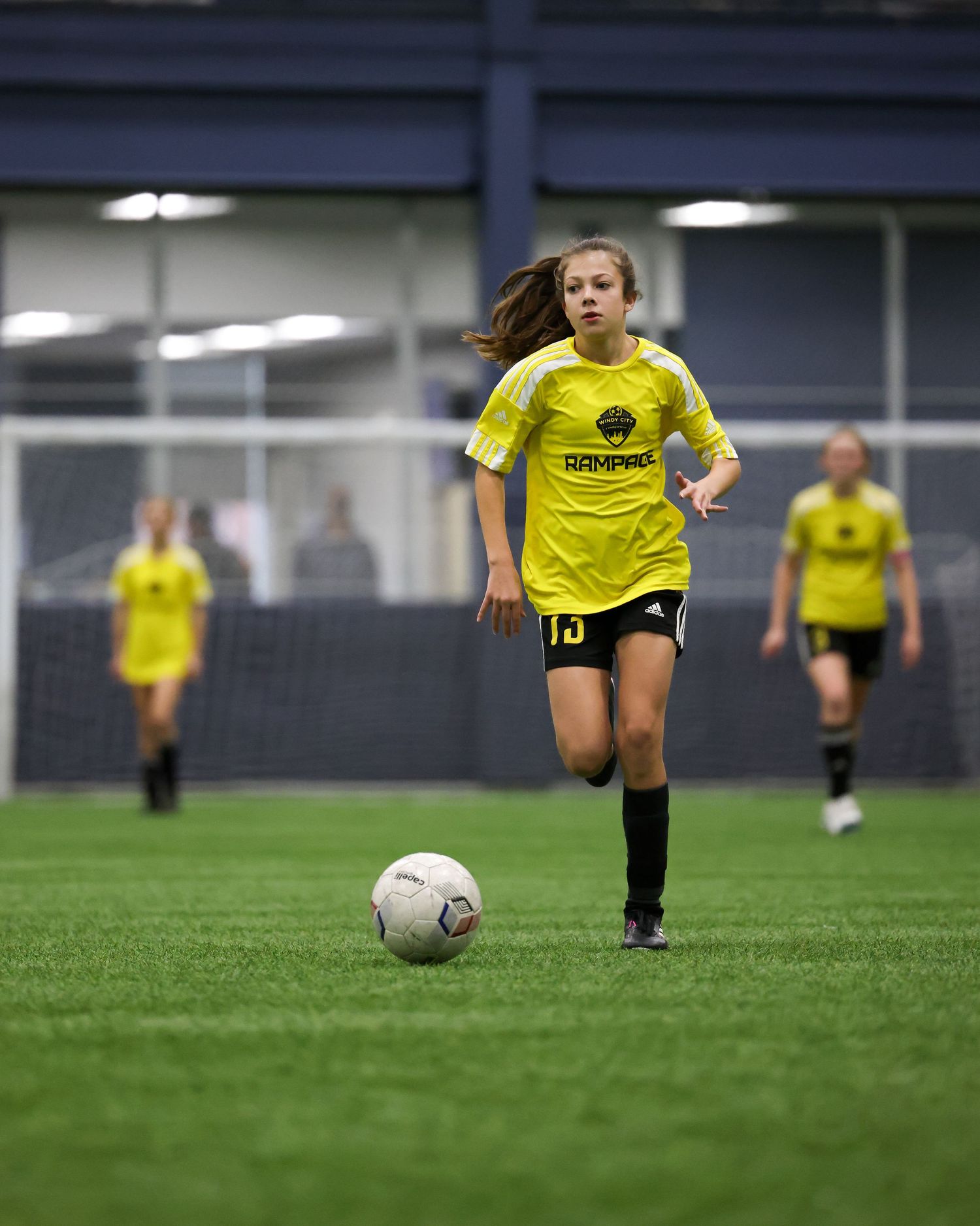 Young soccer players in yellow uniforms practicing ball control drills on an indoor turf field.