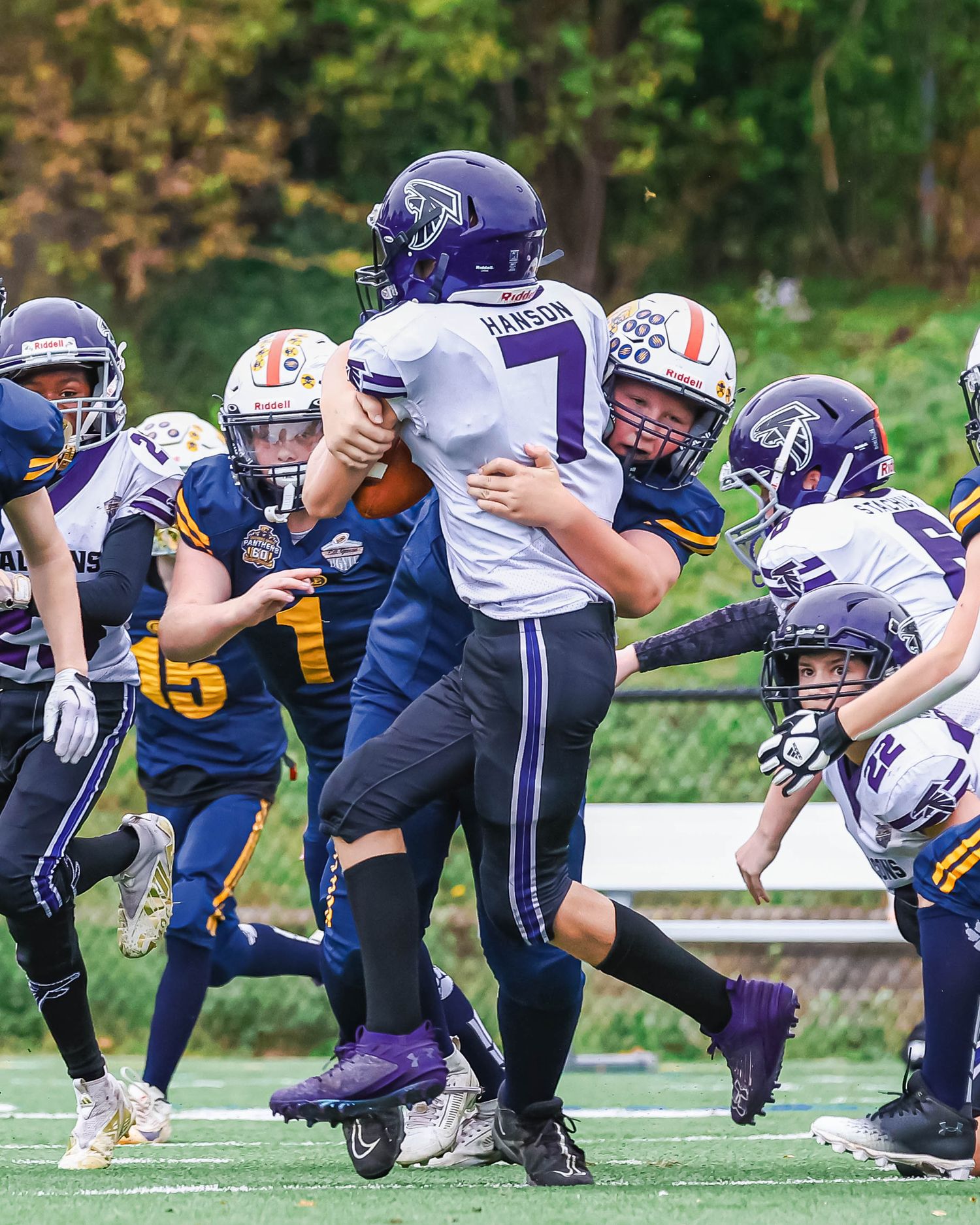 Youth football players in purple and blue uniforms collide during a competitive tackle play on a green field.