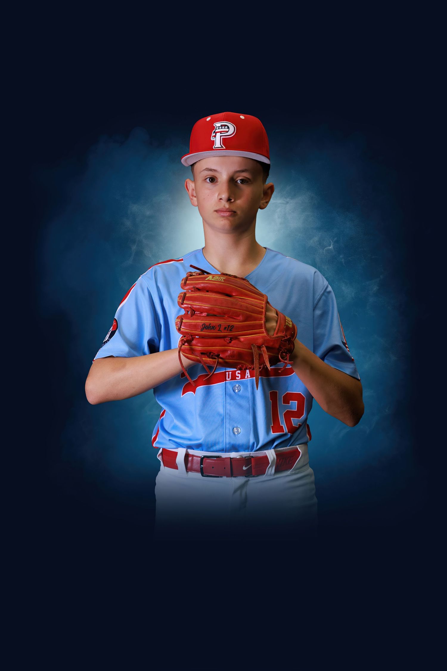 Young baseball pitcher in light blue jersey number 12 and red cap poses with glove on dark blue background.