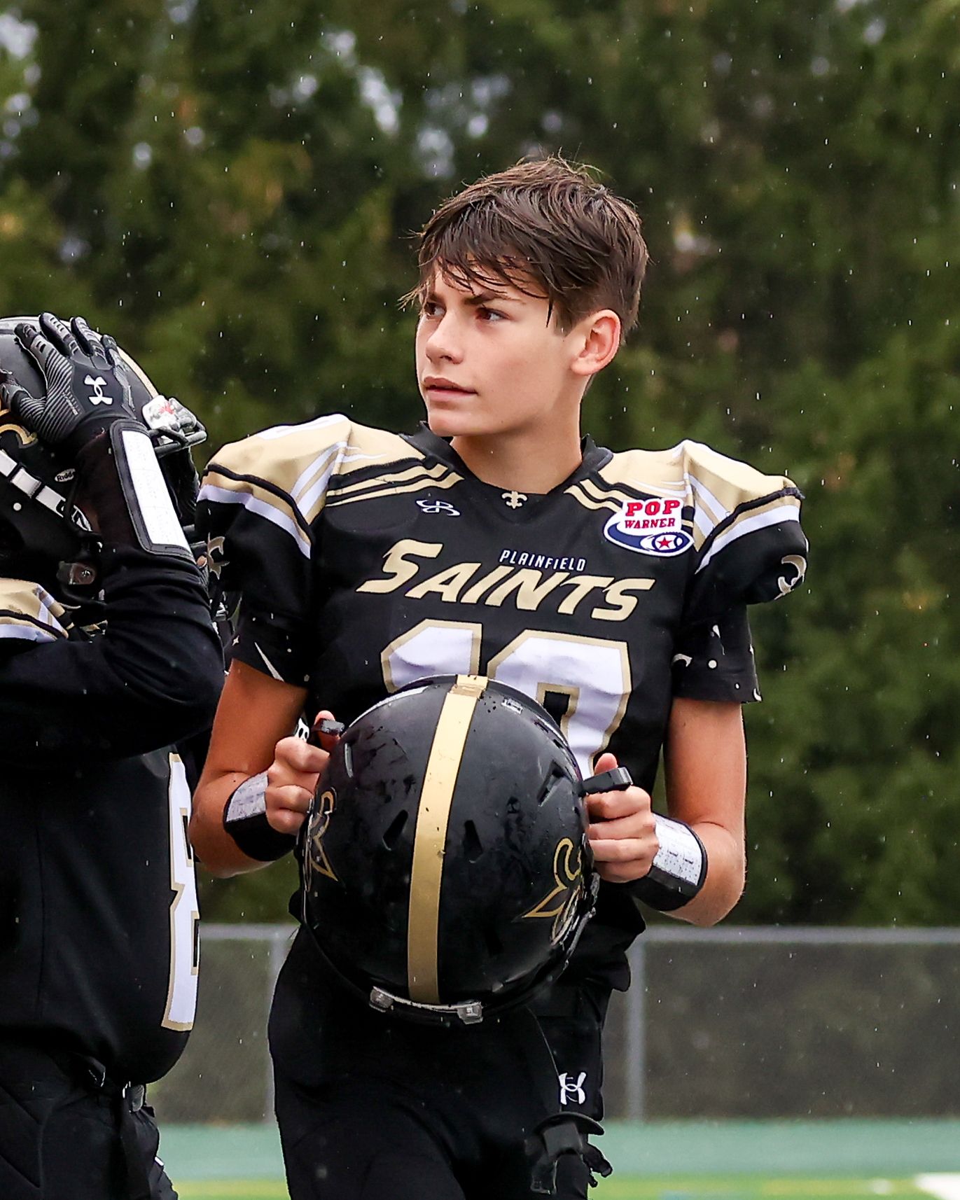 Young Saints football player in black uniform holding helmet on sideline.