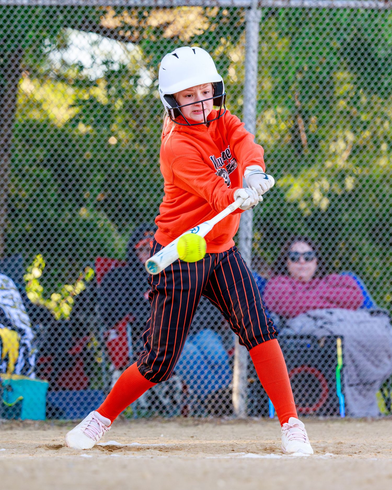Young softball player in orange uniform and white helmet makes contact with a yellow ball during a youth league game.