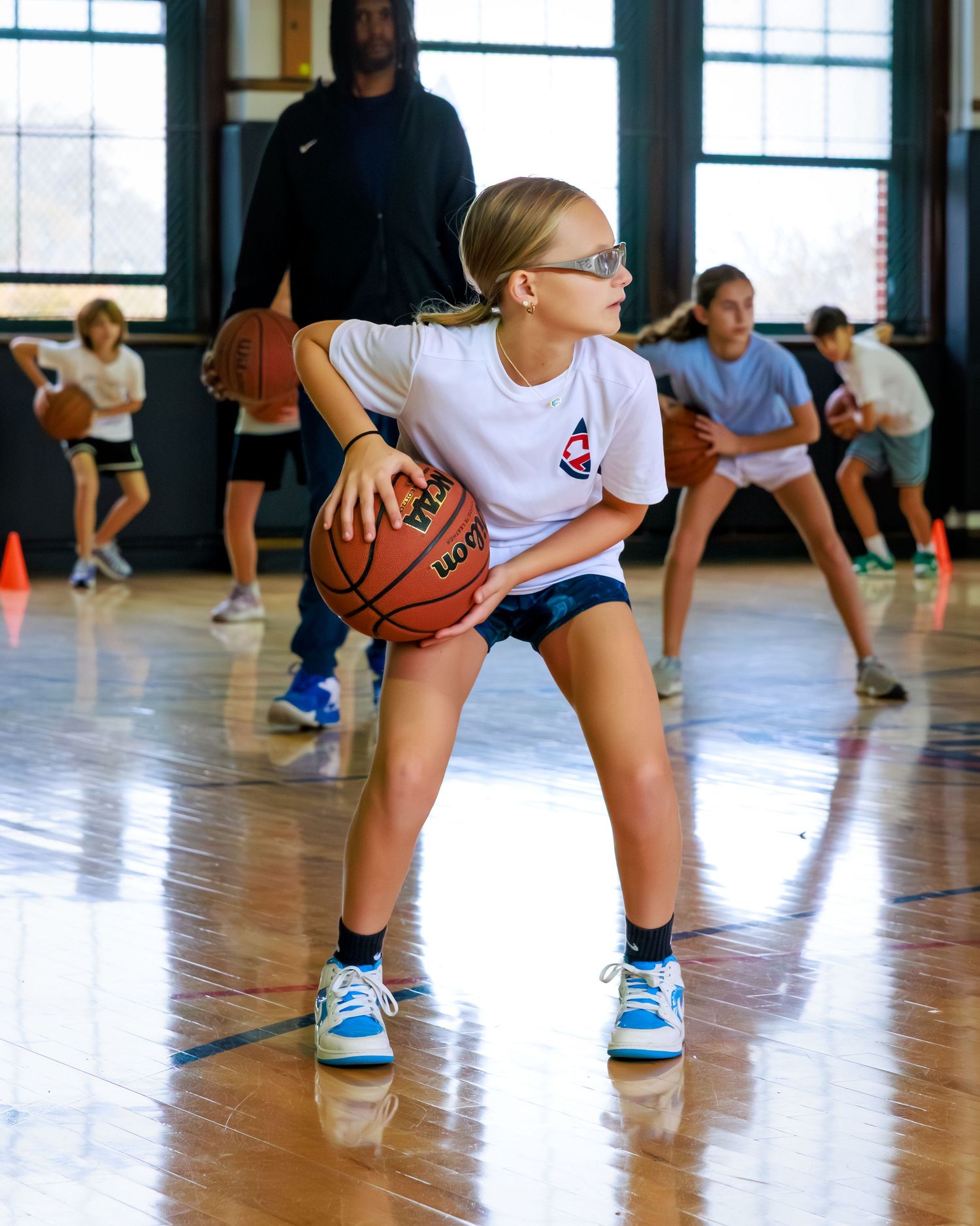 Basketball players practice dribbling drills in a gymnasium with hardwood floors and large windows.