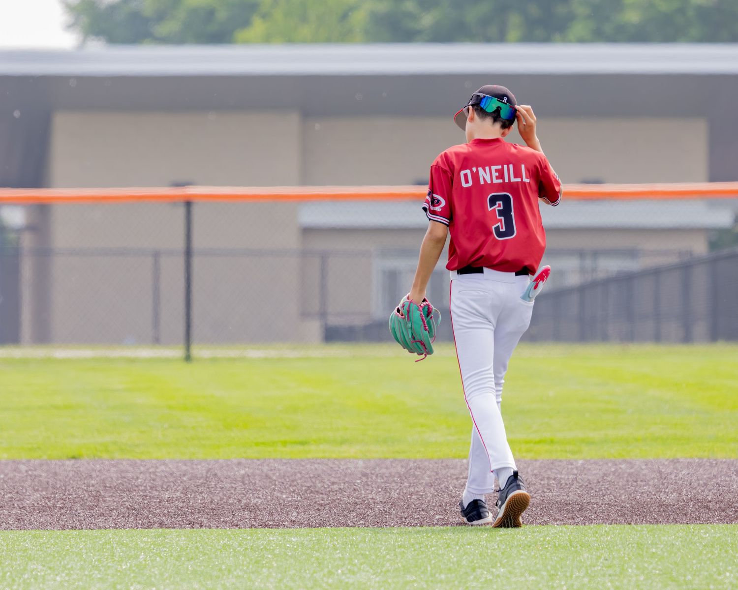 Baseball player in red jersey number 3 walks on field during game, seen from behind with glove in hand.