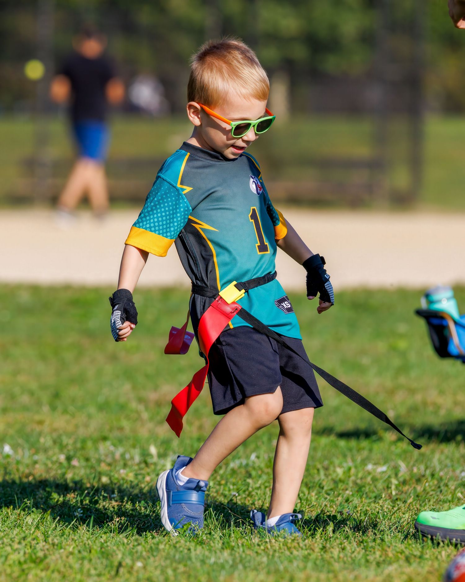 Young boy in flag football jersey, green sunglasses, and colorful flags runs on grass field during youth sports game.