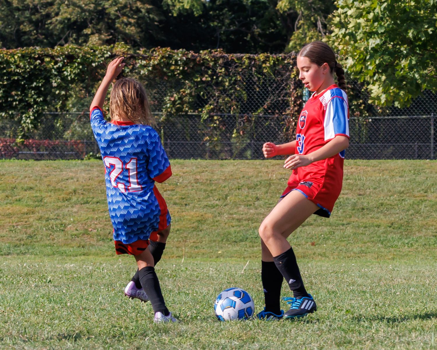 Two girls playing youth soccer, one in blue jersey number 21, other in red, competing for ball on green grass field.