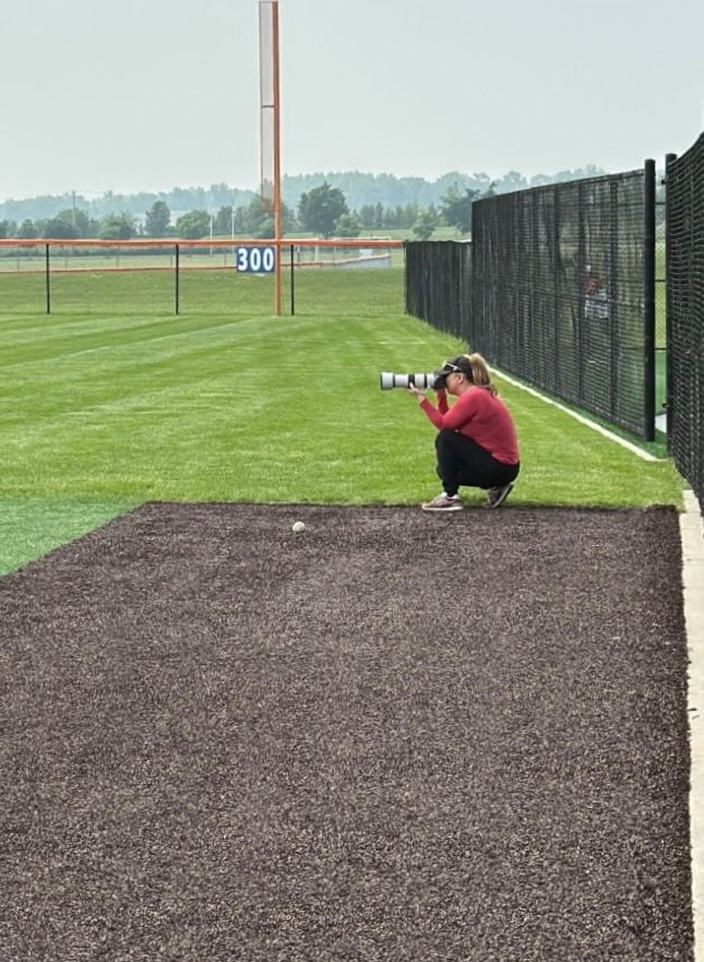 A person crouching down taking with a camera on mulch near a black fence on an athletic field.