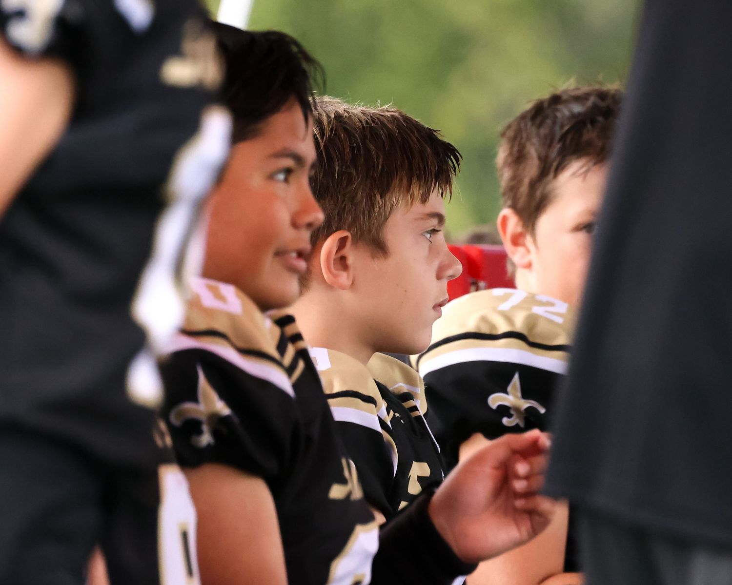 Young boys in black and gold football uniforms with fleur-de-lis logos stand together on the sideline during a game.