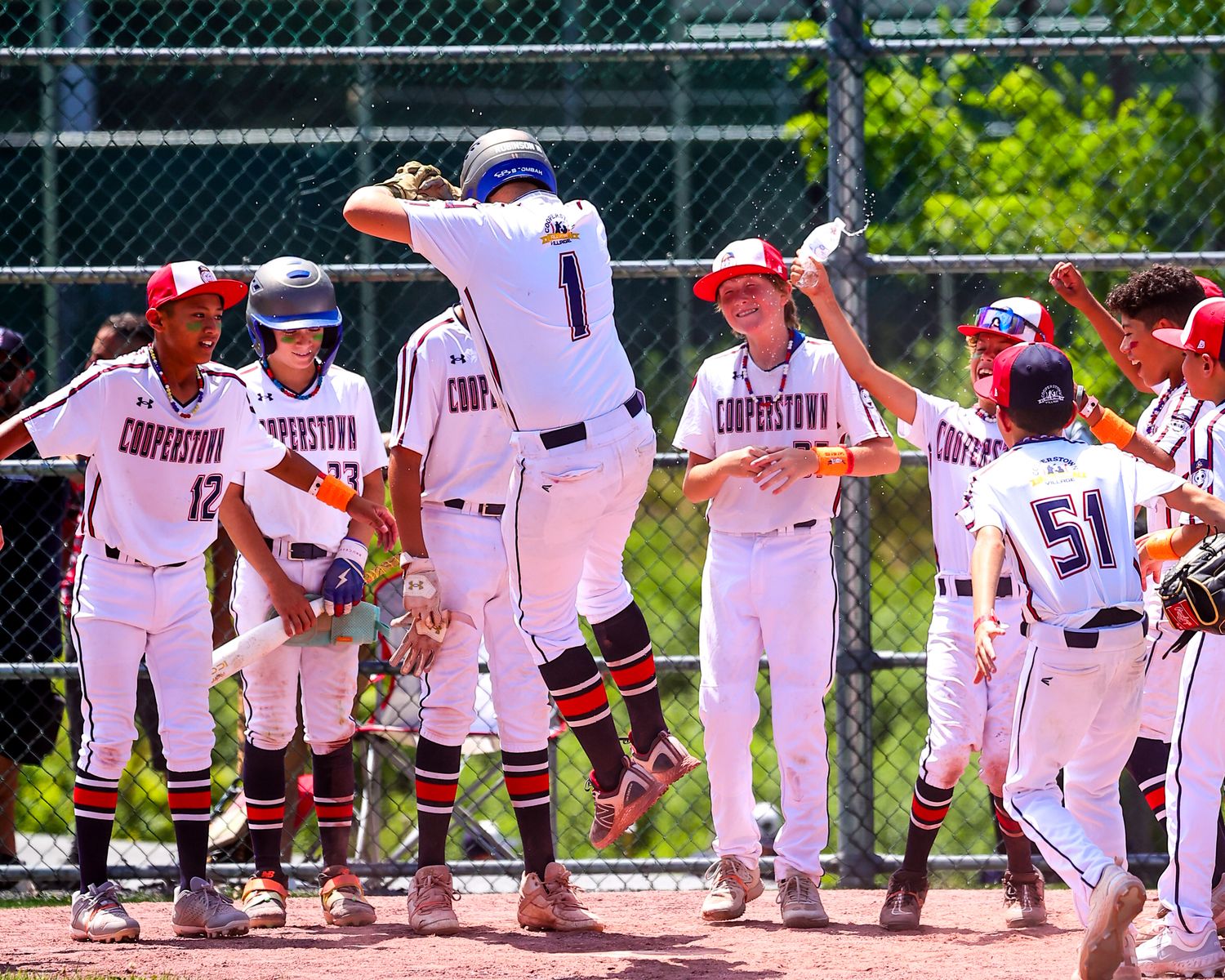 Baseball players in white uniforms line up along a fence during a game on a sunny day at the field.