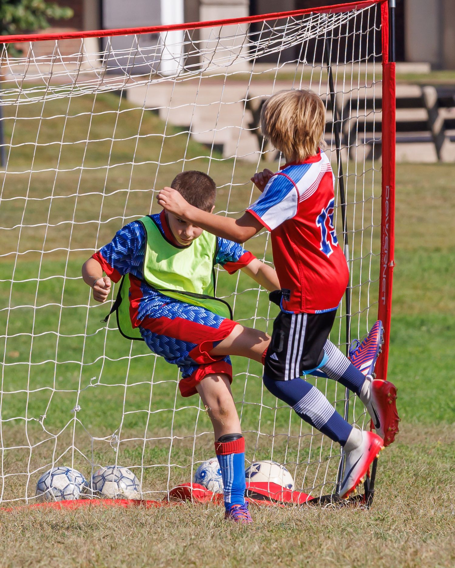 Two young boys in red and blue soccer uniforms compete near a goal net during a youth soccer match outdoors.