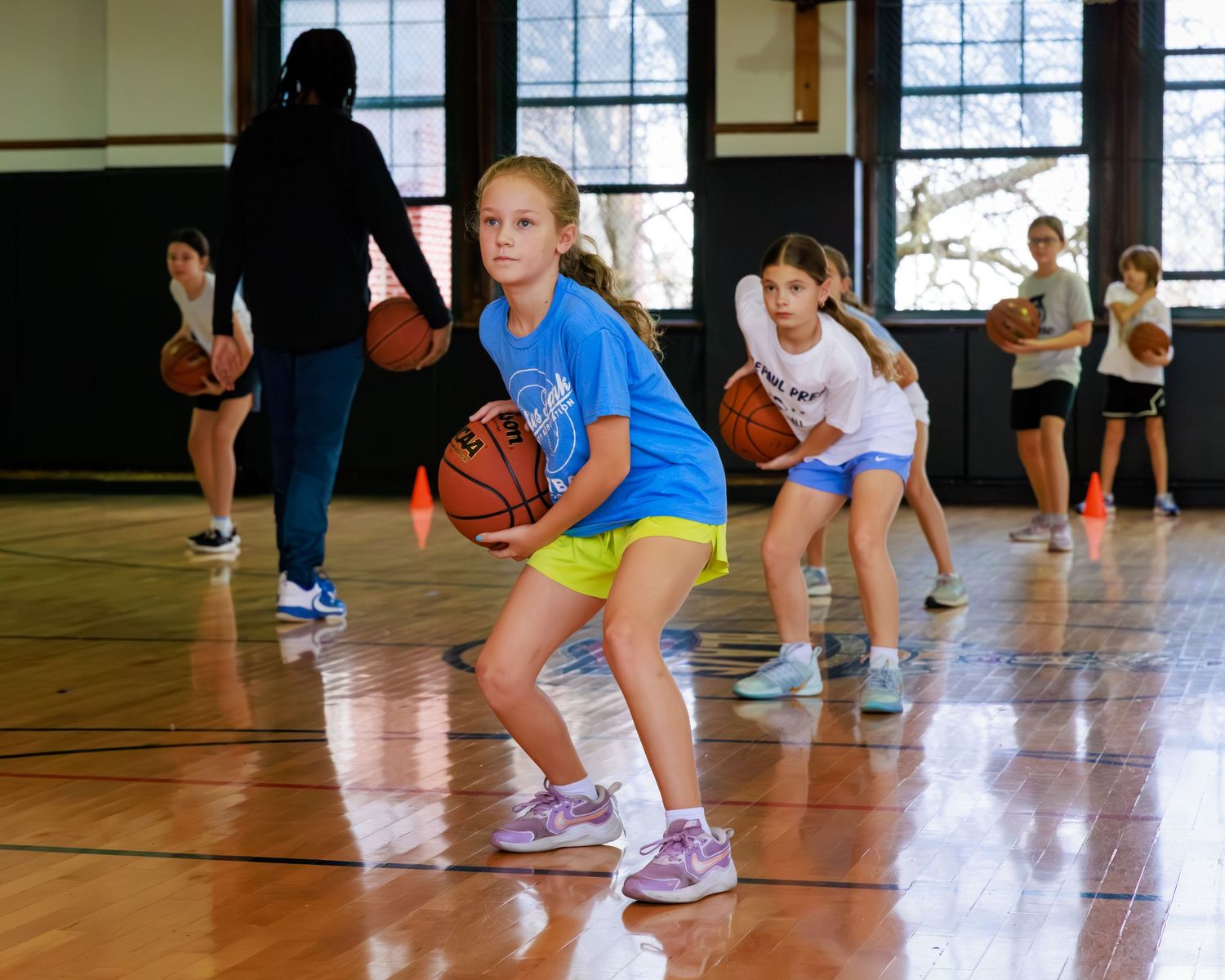 Basketball players practice dribbling drills in a gymnasium with hardwood floors and large windows.