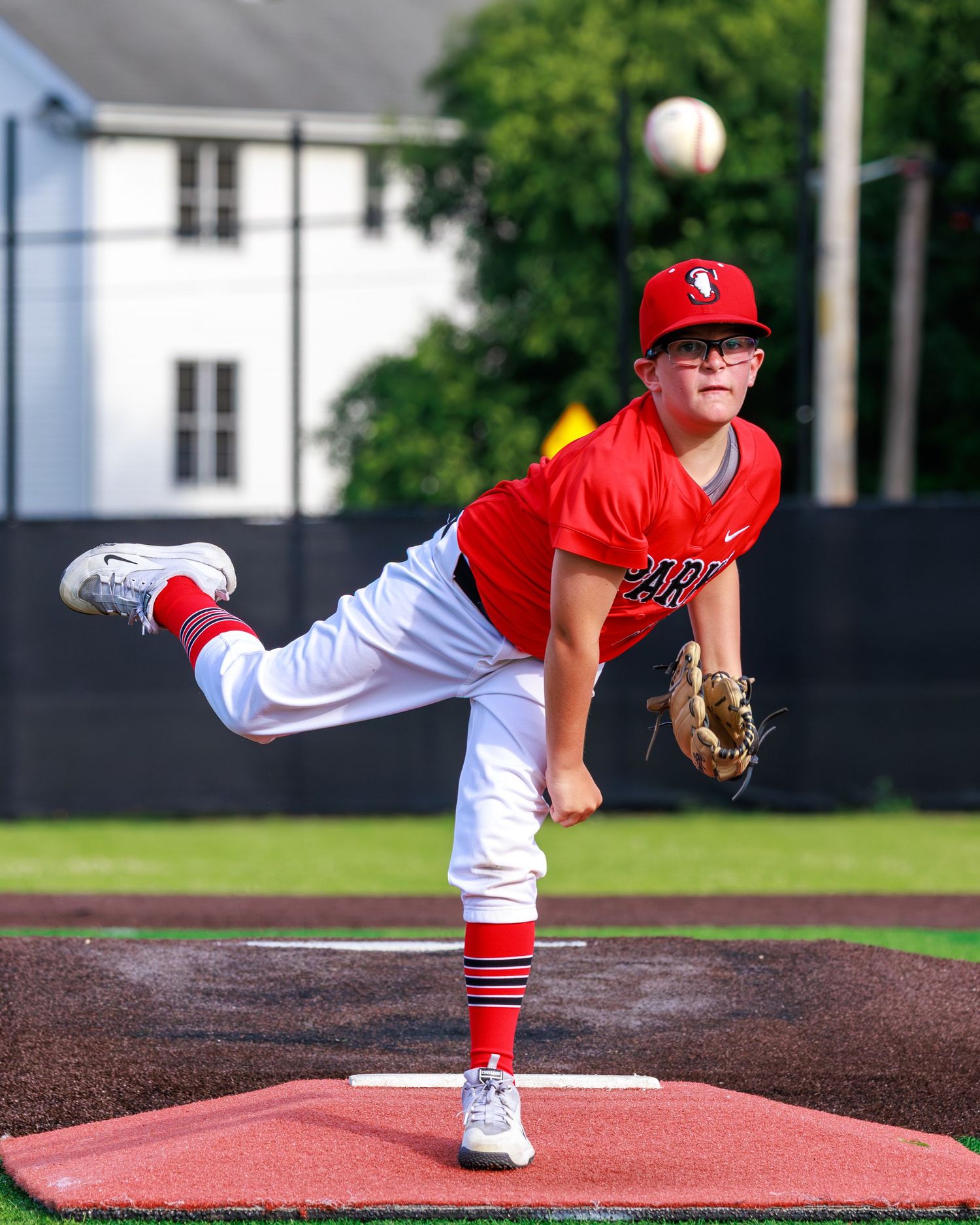 Young baseball pitcher in red uniform mid-throw on mound, ball visible in air, green field background.