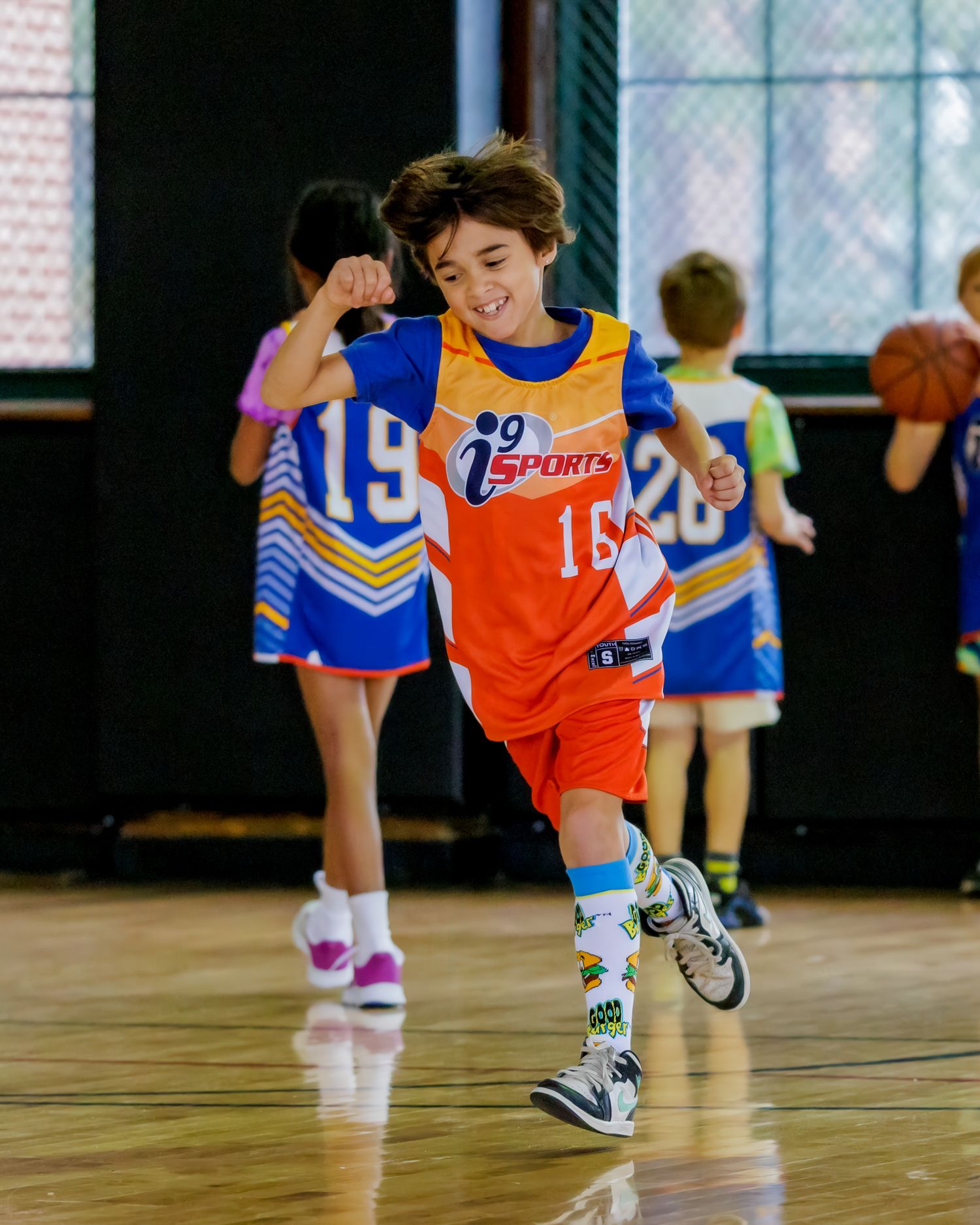 Kids playing basketball in orange and blue jerseys practicing dribbling skills on an indoor court.