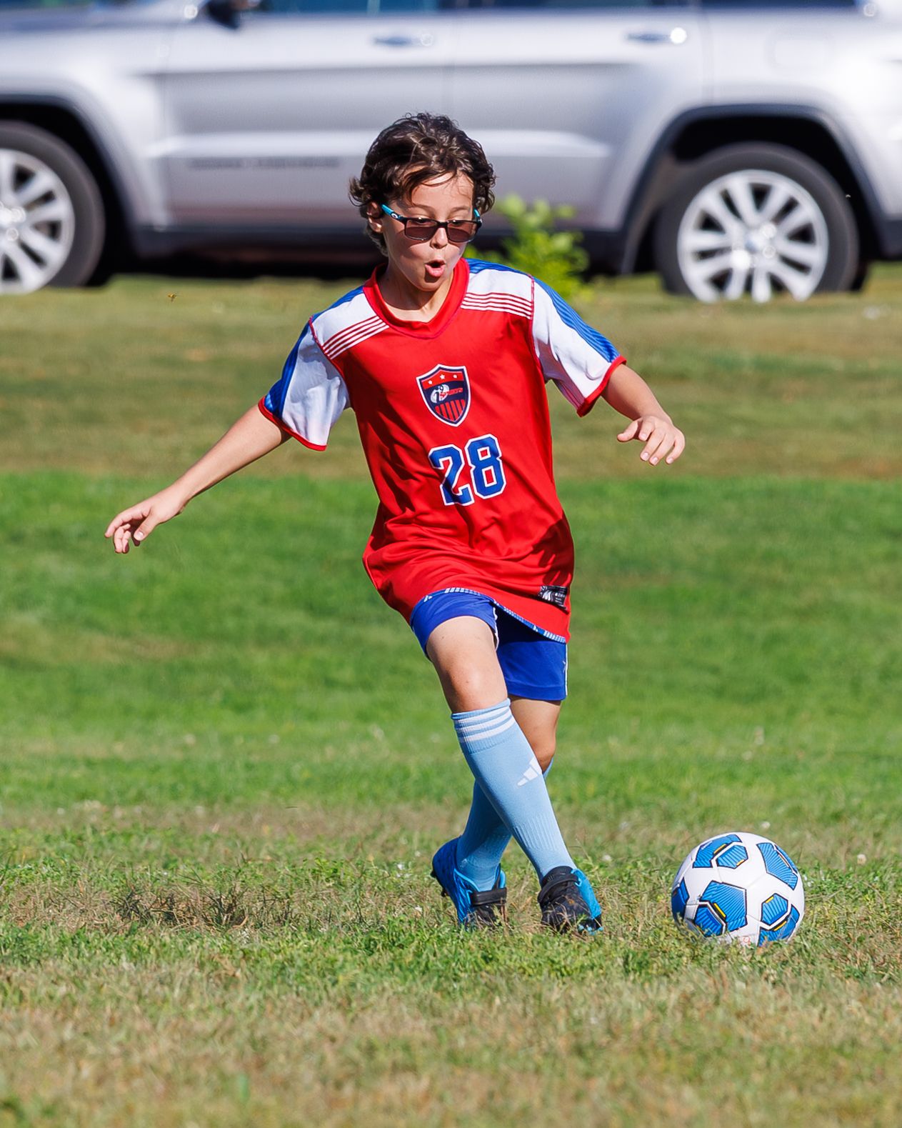 Young soccer player wearing number 28 red jersey and blue socks dribbling a soccer ball on a grass field.