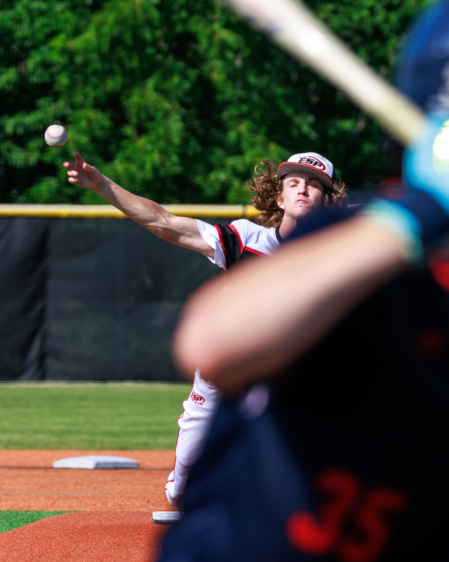 A young baseball pitcher with curly hair winds up to throw a pitch during a sunny outdoor game.