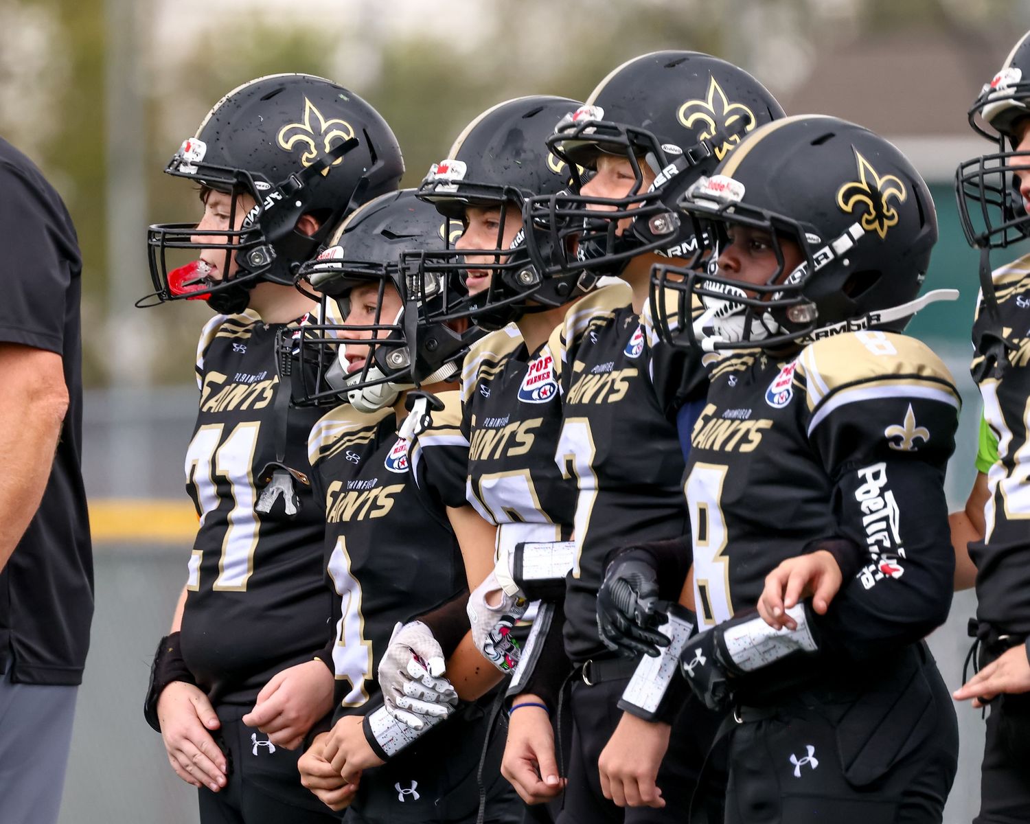 Youth football players in black Saints helmets and uniforms stand on the sideline during a game.