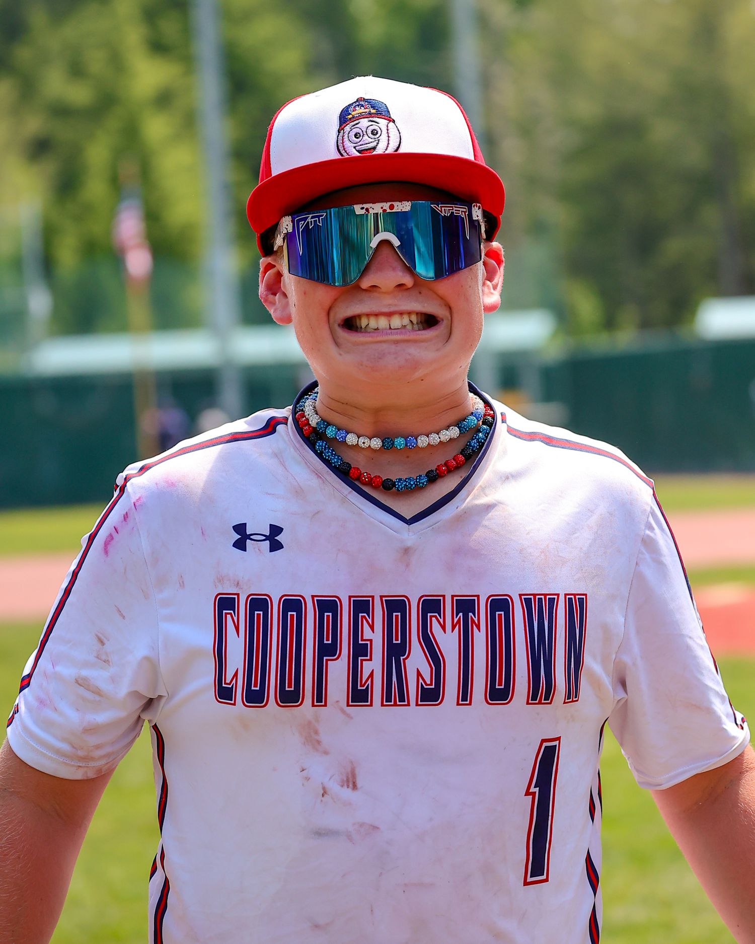 Baseball player in Cooperstown uniform wearing sunglasses and red cap smiles on sunny field.